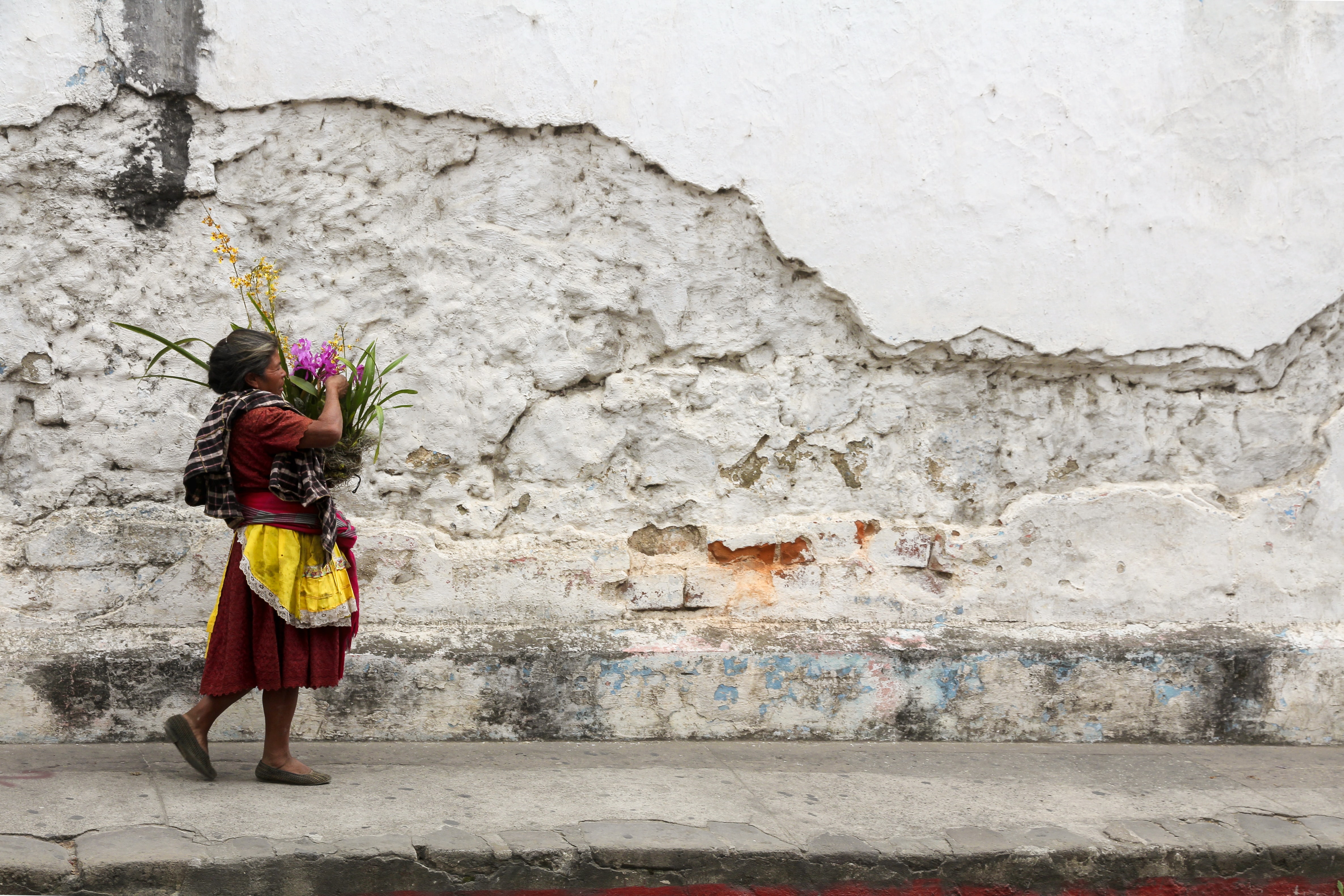 A woman carries a basket of flowers next to the cobblestone streets and crumbling walls of Antigua, Guatemala.